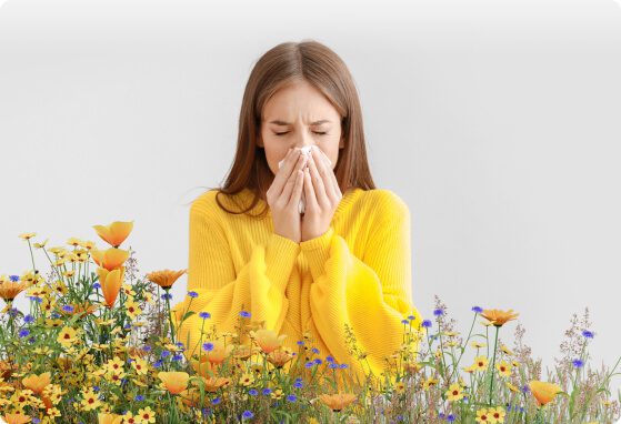 Alergik kobieta w żółtym swetrze zatykająca nos wśród kwiatów, objawy alergii na pyłki.